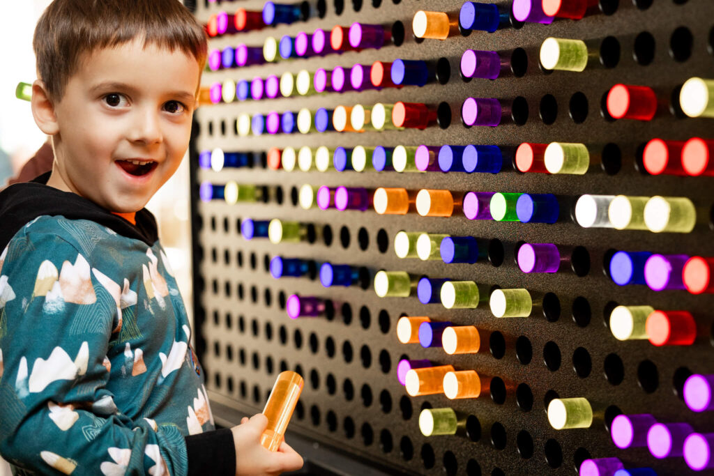A young boy with short brown hair and a playful expression is standing in front of a colorful wall filled with illuminated tubes. He is holding an orange tube in his hand and wearing a patterned hoodie. The wall displays various vibrant colors, including red, blue, green, and purple, creating a visually engaging backdrop.
